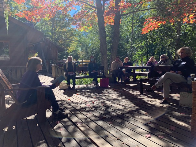 A woman sitting on a wood deck is reading from her work on a beautiful fall day to a group of women