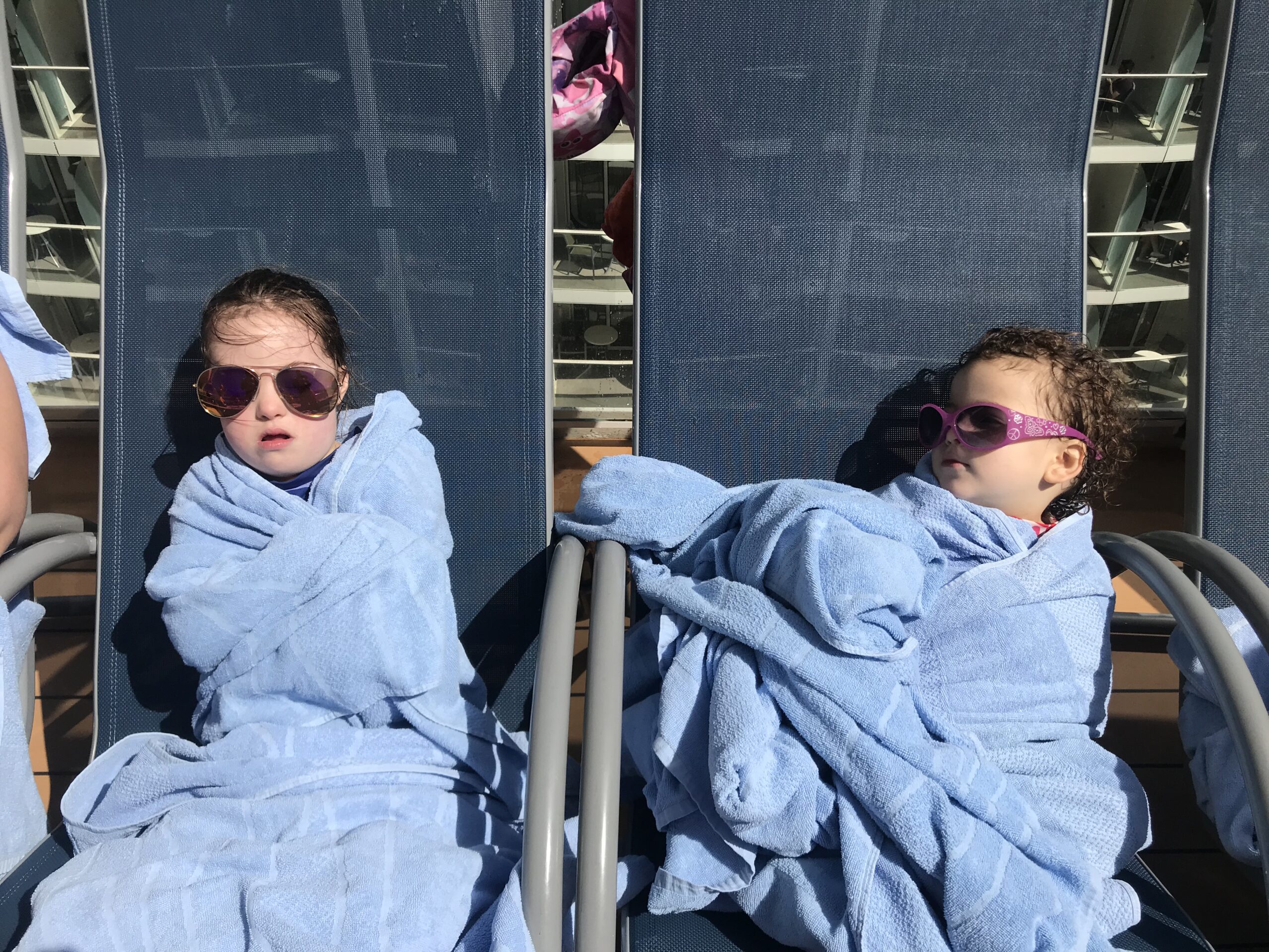 two kids wrapped in blue towels wearing sunglasses sitting in lounger chairs